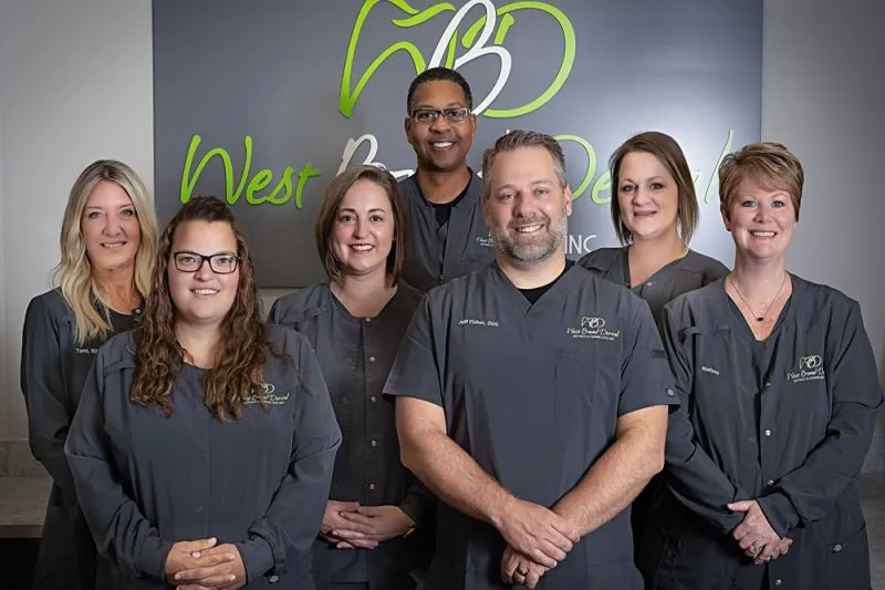 The dental team and staff of West Broad Dental standing together and smiling in front of the office logo in matching gray scrubs.
